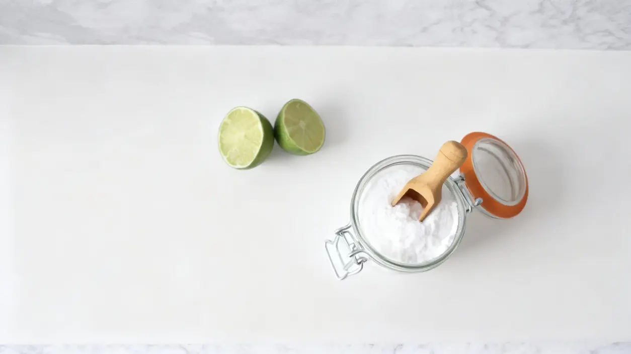 Barattolo di bicarbonato con cucchiaio in legno accanto a due metà di lime su sfondo chiaro