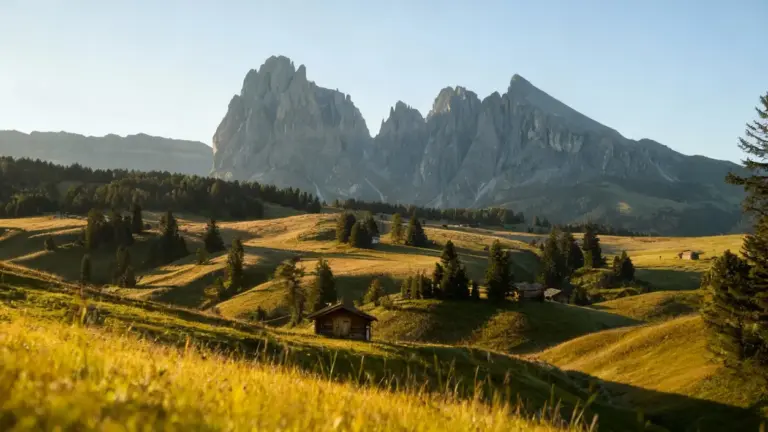 Paesaggio montano con baite in legno tra prati verdi e alberi, sullo sfondo imponenti cime rocciose